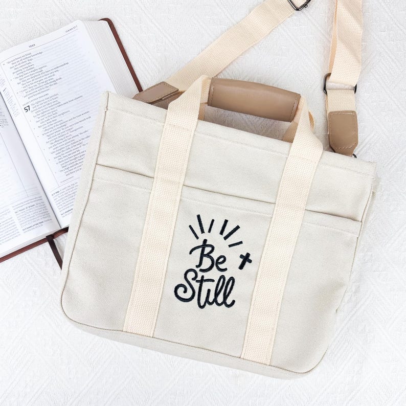 Op de afbeelding: Cr&egrave;me canvas draagtas met de geborduurde tekst "Be Still" in zwart. De tas heeft twee handvatten en een afneembare schouderriem. Op de achtergrond is een open boek zichtbaar.