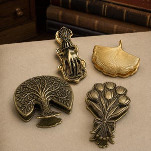 Peut inclure: Quatre pinces en métal avec une finition en laiton antique. Une pince représente une main avec une bague, une autre une feuille de ginkgo, une troisième un arbre aux branches étendues et la dernière un bouquet de tulipes noué par un ruban.