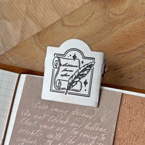 May include: A silver-toned metal page clip featuring an engraved design of a rolled parchment scroll and a quill pen. The central graphic includes the text 'Letter from afar' surrounded by small star accents. The clip is attached to a piece of paper.
