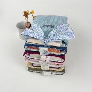 May include: A stack of colorful, corduroy book covers, each with a different color and pattern. The top cover is light blue with the name "Michelle" embroidered on it. A floral bow ties the stack together. A small vase with dried flowers is in the background.