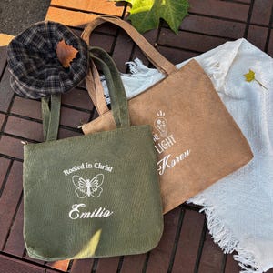 May include: Two corduroy tote bags, one olive green and one tan, with long handles. The green bag has the text "Rooted in Christ" and "Emilia" with a butterfly graphic. The tan bag has the text "THE LIGHT" and "Karen". A plaid hat is on the left.
