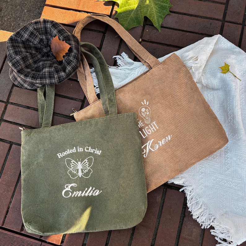 May include: Two corduroy tote bags, one olive green and one tan, with long handles. The green bag has the text "Rooted in Christ" and "Emilia" with a butterfly graphic. The tan bag has the text "THE LIGHT" and "Karen". A plaid hat is on the left.