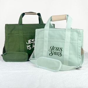 Op de afbeelding: Twee canvas draagtassen, &eacute;&eacute;n in olijfgroen en de andere in lichtblauw. Elke tas heeft een bijpassend afneembaar etui en is voorzien van de tekst "JESUS IS KING" of "JESUS SAVES" in witte letters. De tassen hebben beige handvatten.