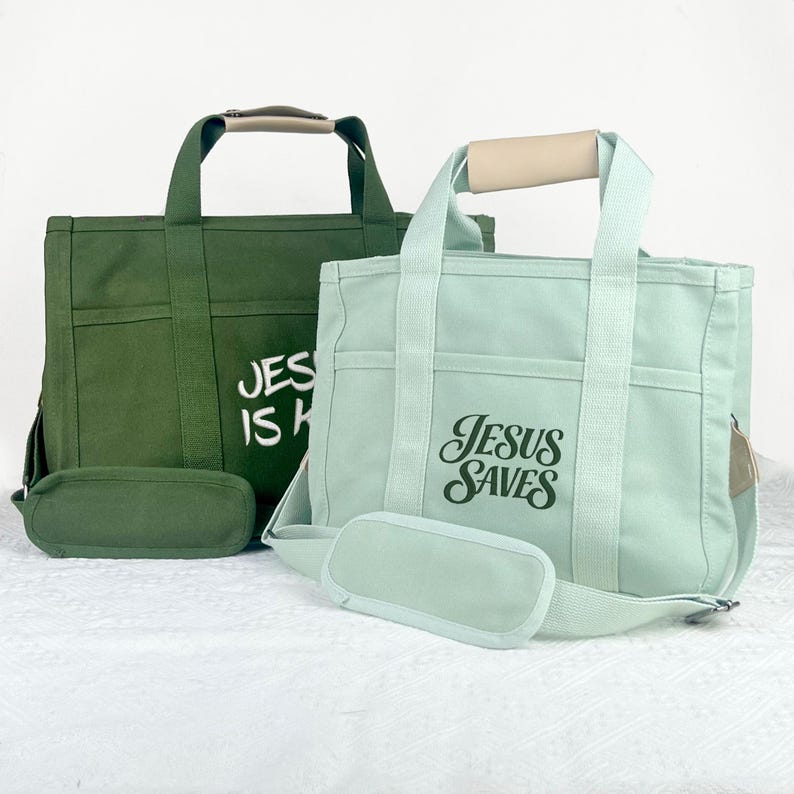 Op de afbeelding: Twee canvas draagtassen, &eacute;&eacute;n in olijfgroen en de andere in lichtblauw. Elke tas heeft een bijpassend afneembaar etui en is voorzien van de tekst "JESUS IS KING" of "JESUS SAVES" in witte letters. De tassen hebben beige handvatten.