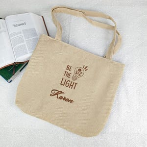 May include: Beige corduroy tote bag with the text "BE THE LIGHT" and the name "Karen" printed in brown. The bag features a lightbulb design with a flower inside. The bag has two beige handles and is next to an open book.