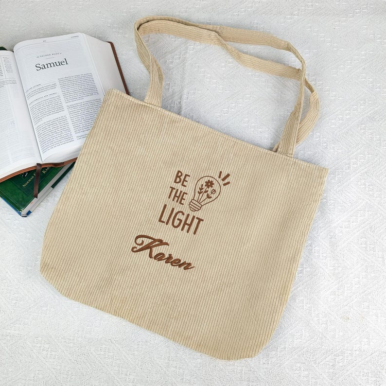 May include: Beige corduroy tote bag with the text "BE THE LIGHT" and the name "Karen" printed in brown. The bag features a lightbulb design with a flower inside. The bag has two beige handles and is next to an open book.