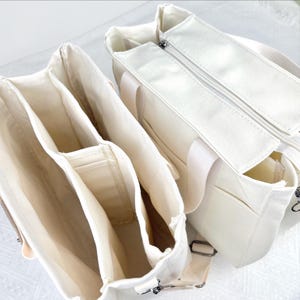 Op de afbeelding: Twee off-white canvas draagtassen. E&eacute;n tas is open en toont meerdere binnenvakken. De andere tas is gesloten met een rits. Beide tassen hebben handvatten en verstelbare schouderbanden. De tassen zijn ontworpen om spullen te dragen.