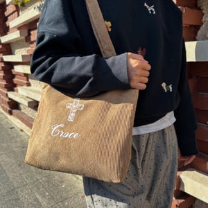 May include: A tan corduroy tote bag with a shoulder strap. The bag features an embroidered cross design above the word "Grace" in white. The bag is rectangular and has vertical corduroy ridges.