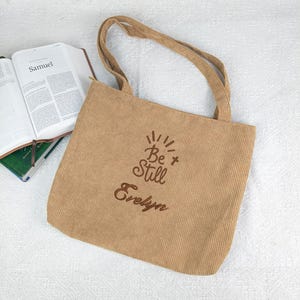 May include: A tan corduroy tote bag with the embroidered text "Be Still Evelyn" and a cross design. The bag has two handles and a zipper closure. An open book with the title "Samuel" is visible in the background.