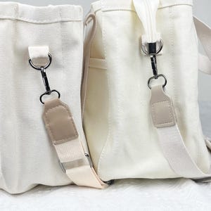 Op de afbeelding: Twee off-white canvas draagtassen met cr&egrave;mekleurige bandjes en zilverkleurige metalen hardware. E&eacute;n tas heeft een zijvak. De bandjes zijn bevestigd met metalen sluitingen en beige leren accenten.