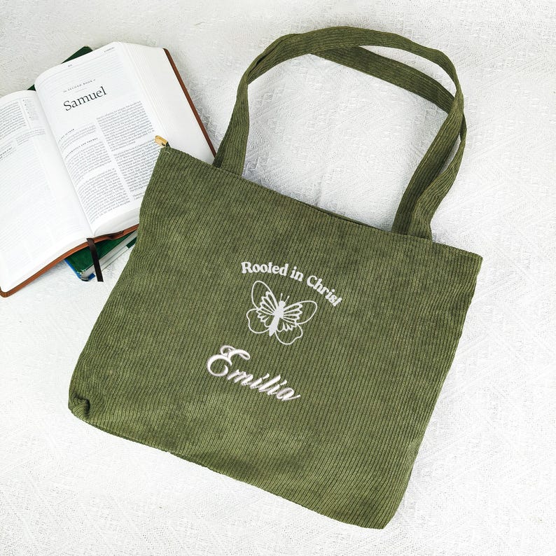 May include: Olive green corduroy tote bag with long handles. The bag features white embroidered text that reads "Rooted in Christ", a butterfly design, and the name "Emilia". An open book is visible in the background.