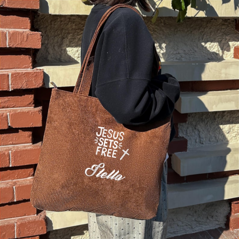 May include: A brown corduroy tote bag with the text "JESUS SETS FREE" and a cross graphic in white. The name "Stella" is embroidered below. The bag has a zipper closure and long straps.