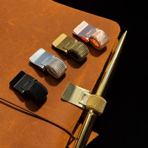 May include: A collection of five metal pen holders in black, silver, bronze, copper, and gold finishes. Each holder features a flat clip base and a coiled spring loop designed to secure a writing instrument, with one gold pen inserted.