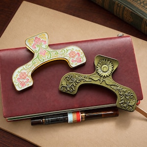 May include: Two metal hair clips with ornate designs. One clip features white enamel with pink and green floral patterns. The second clip is made of antique brass-toned metal and displays a sunflower motif with intricate scrolling vine details.