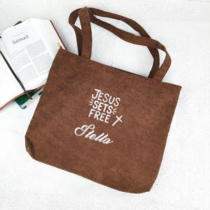 May include: A brown corduroy tote bag with the text "JESUS SETS FREE Stella" embroidered in white. The bag has two handles and a zipper closure. An open book with the title "Samuel" is visible in the background.
