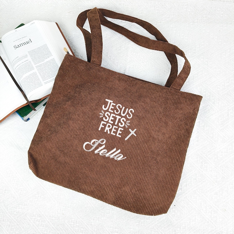 May include: A brown corduroy tote bag with the text "JESUS SETS FREE Stella" embroidered in white. The bag has two handles and a zipper closure. An open book with the title "Samuel" is visible in the background.