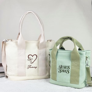 May include: A large off-white canvas tote bag with a brown heart and cross graphic and the name Jenny, alongside a smaller sage green canvas tote bag featuring the dark green text Jesus Saves. Both bags have sturdy handles and detachable straps.