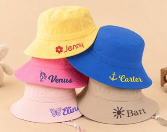 May include: A stack of five colorful bucket hats in yellow, pink, blue, and tan. Each hat has a different name embroidered on it in a contrasting color, such as "Jenny" and "Carter". The hats are made of a soft fabric and have a brim.
