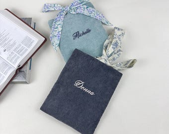 May include: Two corduroy book covers, one light blue and one navy, with embroidered names "Michelle" and "Donna". Each cover has a floral patterned ribbon tied in a bow. An open book is visible in the background.
