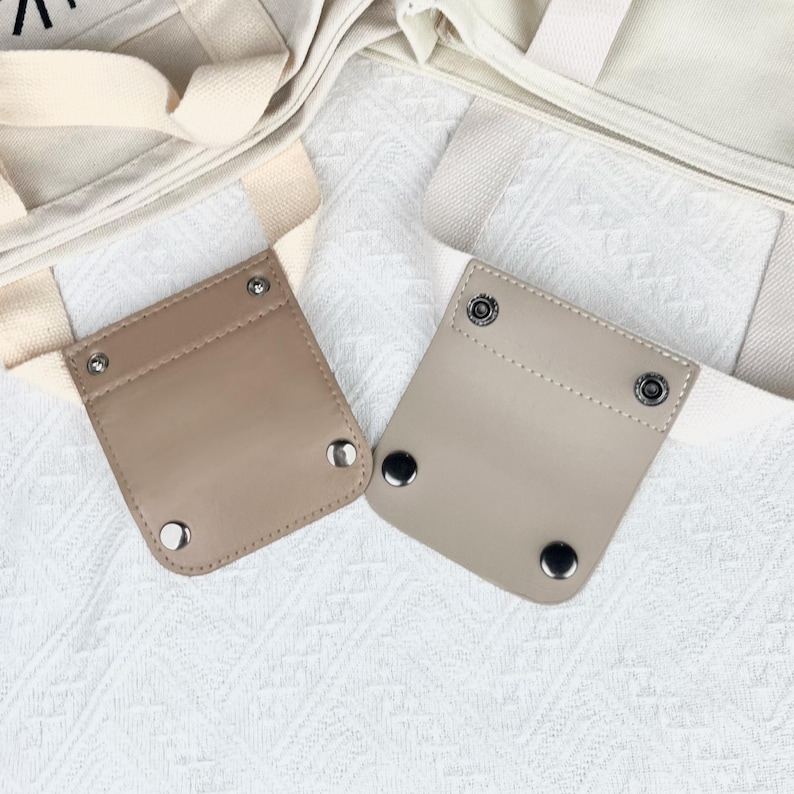 Op de afbeelding: Twee beige leren tasriemhouders met metalen druksluitingen. De houders zijn rechthoekig met afgeronde hoeken en zijn bevestigd aan witte canvas draagtasriemen. De achtergrond is een witte, getextureerde stof.