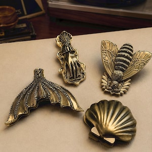 May include: A set of four antique brass-toned metal clips featuring distinct designs: a mermaid tail, a hand with a cuff, a detailed bee with spread wings, and a scalloped seashell. Each clip has a metallic finish with dark recessed detailing.