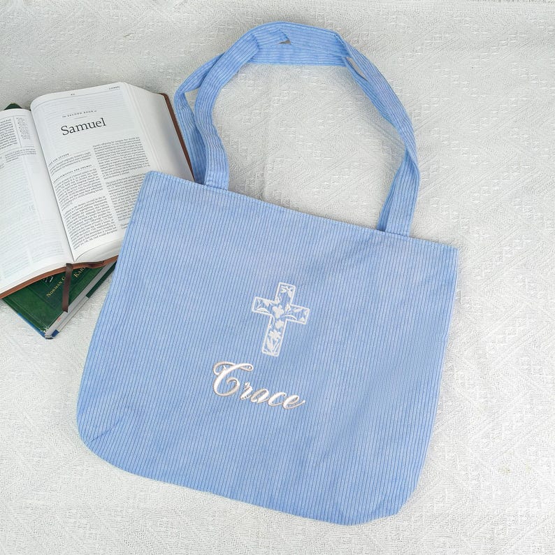 May include: A light blue corduroy tote bag with a white embroidered cross and the word "Grace". The bag has matching handles and is next to an open book with the title "Samuel".