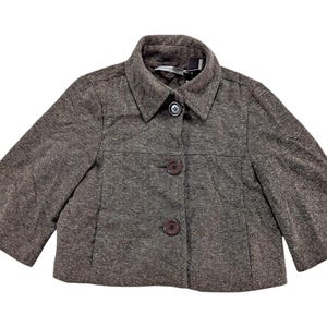 May include: A brown cropped jacket with three-quarter sleeves and a collared neckline. The jacket has three dark brown buttons down the front and a textured fabric. The jacket is a neutral color and could be worn with many outfits.