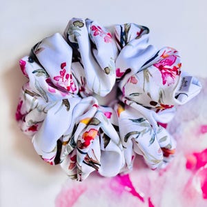 May include: A white scrunchie with a floral pattern featuring pink, red, and green watercolor-style flowers. The fabric is gathered and ruffled, creating a voluminous look. The scrunchie is a hair accessory.