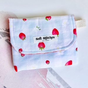 May include: A small, light blue fabric wallet with a flap closure. The wallet is decorated with a pattern of red strawberries and white flowers. The label on the wallet reads "Soft Stitches by Tara."