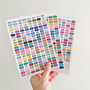 May include: Three rectangular sticker sheets with rounded corners, featuring rows of numbered labels in various colors. The labels have white text on a colored background. The sheets are held by a hand.