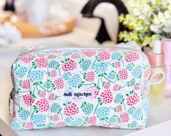 May include: A white cosmetic bag featuring a floral design with pink and blue flowers and green stems. The bag has a zip closure and a small loop. The text "soft stitches" is printed on a white label.