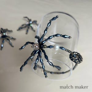 Könnte beinhalten: Dunkelblaue, perlenbesetzte Spinnendekoration mit Perlenakzent und dekorativem, gedrehtem Schwanz. Die Spinne befindet sich in der Nähe eines klaren Glases. Der Text "match maker" ist unten im Bild zu sehen.