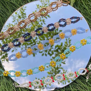 Può includere: Una collezione di braccialetti di perline in vari colori, tra cui oro, blu navy, azzurro e bianco. Alcuni braccialetti presentano disegni floreali con accenti gialli e bianchi. I braccialetti sono esposti su una superficie riflettente.