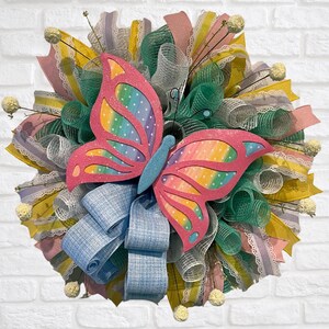 May include: A colorful spring wreath featuring a large pink butterfly with rainbow wings and blue accents. The wreath is made of various ribbons in shades of pink, yellow, green, and blue, with small floral accents. Perfect for spring or Easter decor.