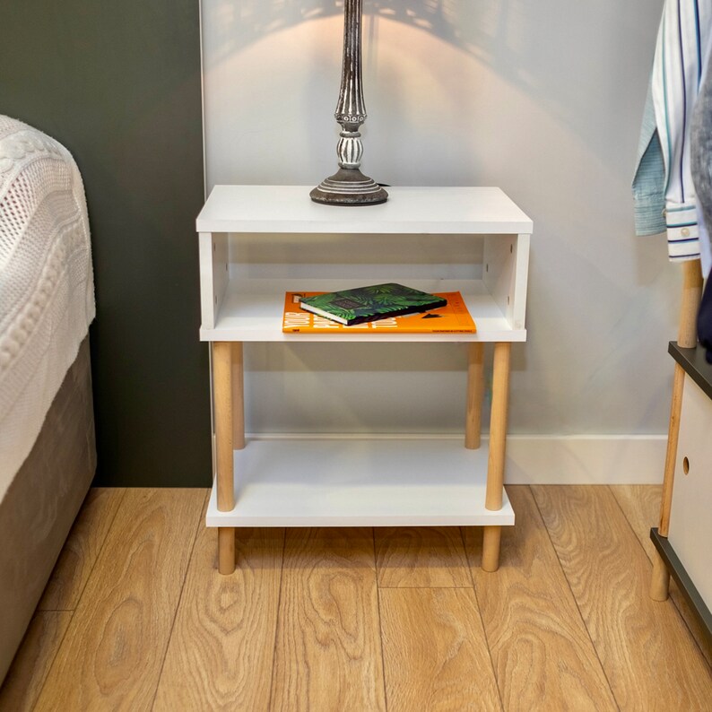 May include: White wooden nightstand with two shelves and wooden legs. A book with a green and brown cover is on the top shelf.