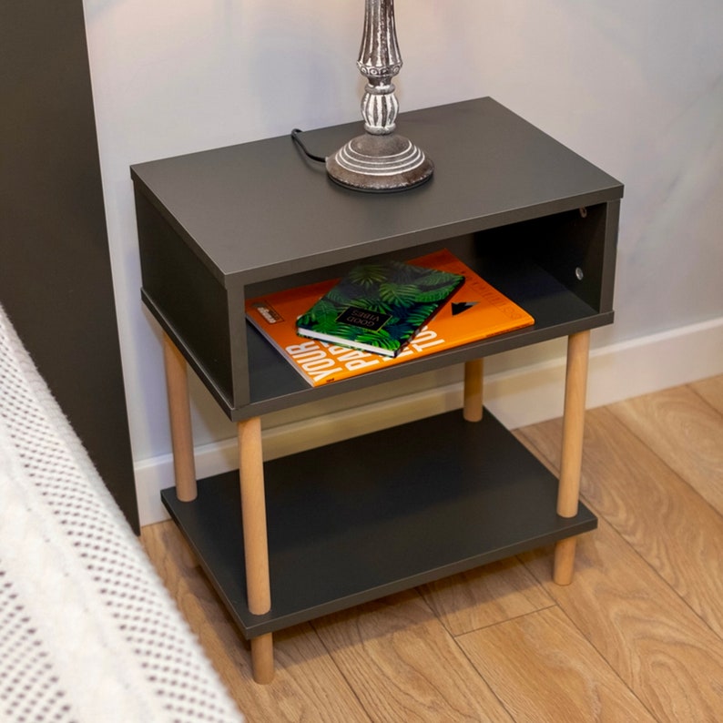 May include: A dark gray nightstand with two shelves and wooden legs. The top shelf has a book with a green and white cover and a book with an orange cover. The nightstand is next to a white bed.