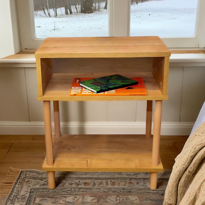 May include: A light wood end table with two shelves and turned legs. A book with a green cover is on the top shelf.