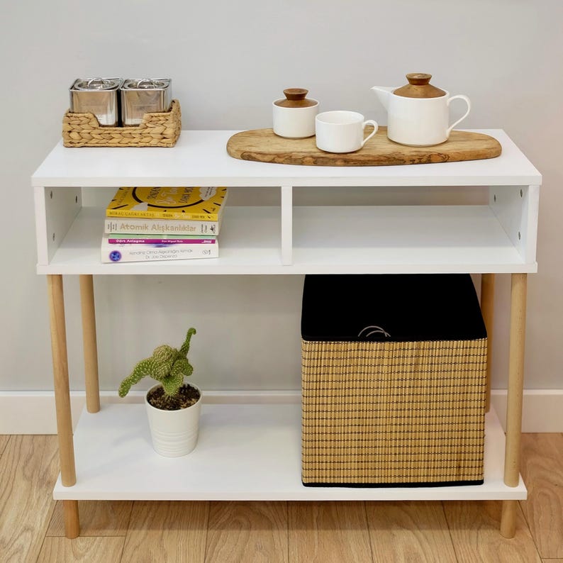 May include: White console table with two shelves, wooden legs, and a woven basket on the bottom shelf. The top shelf has a wooden serving tray with a teapot, sugar bowl, and a cup. There are three small metal canisters on the top shelf.