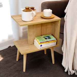 Pode incluir: Uma mesa de apoio de madeira clara com uma prateleira e duas prateleiras inferiores. A mesa tem quatro pernas de madeira e é decorada com um bule branco e uma xícara na prateleira superior. Há três livros na prateleira inferior.