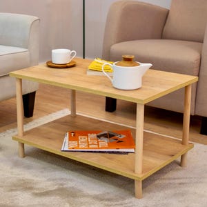 Puede incluir: Una mesa de café de madera clara con dos estantes. La mesa tiene un acabado de madera clara y patas de madera natural. Una tetera blanca y una taza blanca están en el estante superior. Un libro y una revista están en el estante inferior.