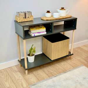 May include: A dark gray console table with two wooden legs on each side. The table has two shelves, one with a woven basket and the other with books. There is a wooden tray on the top shelf with three small ceramic containers and a teapot.