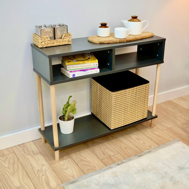 May include: A dark gray console table with two wooden legs on each side. The table has two shelves, one with a woven basket and the other with books. There is a wooden tray on the top shelf with three small ceramic containers and a teapot.
