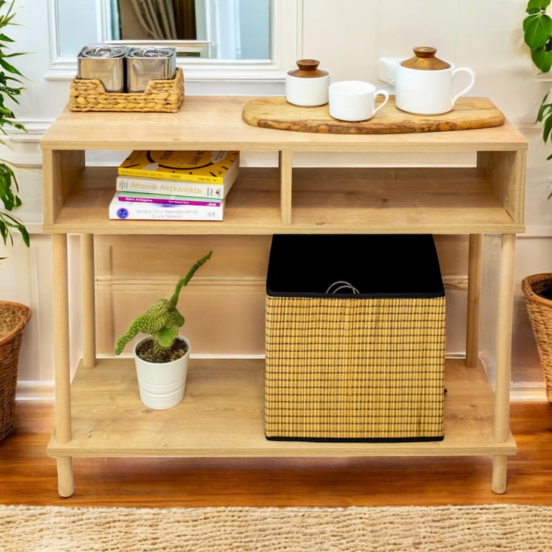 May include: A light wood console table with two shelves and a woven basket. The table has a wooden top and a woven basket on the bottom shelf. There are books and a potted plant on the top shelf.