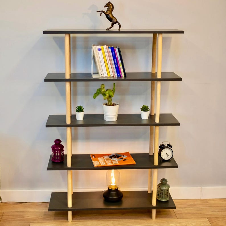 Op de afbeelding: Een zwarte boekenkast met vier planken en houten poten. De planken zijn versierd met boeken, planten, een kleine lamp en een klok.