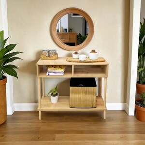 May include: A light wood console table with two shelves, a round mirror, and a woven basket. The table has a wooden top and a woven tray with two white ceramic teapots.