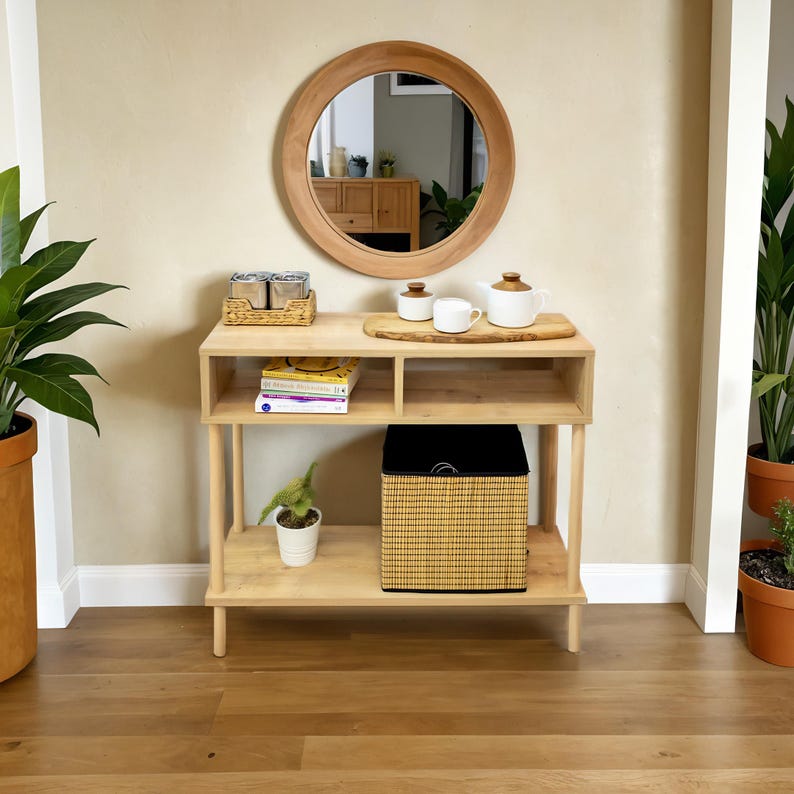 May include: A light wood console table with two shelves, a round mirror, and a woven basket. The table has a wooden top and a woven tray with two white ceramic teapots.