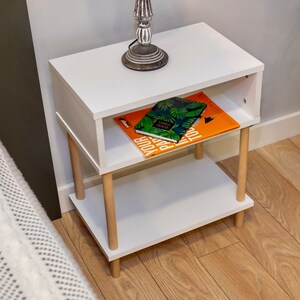 May include: White bedside table with two shelves and wooden legs. The top shelf has a lamp and two books, one with a green and white cover and the other with an orange cover. The text on the book with the green and white cover reads "Serbia Good Vibes".