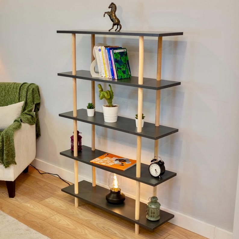 Op de afbeelding: Een boekenkast met vijf planken, een donkergrijze afwerking en natuurlijke houten palen. De planken zijn versierd met boeken, planten, een kleine lamp en een klok.