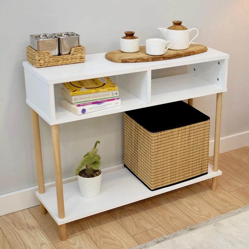 May include: White console table with two shelves and wooden legs. The top shelf has a wooden tray with two small silver containers and a small potted succulent plant. The bottom shelf has a woven basket with a black liner.
