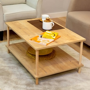 Op de afbeelding: Een lichte houten salontafel met een lager plank. De tafel heeft een natuurlijke houten afwerking en wordt ondersteund door vier slanke poten. Twee boeken en een witte mok staan op de bovenste plank.